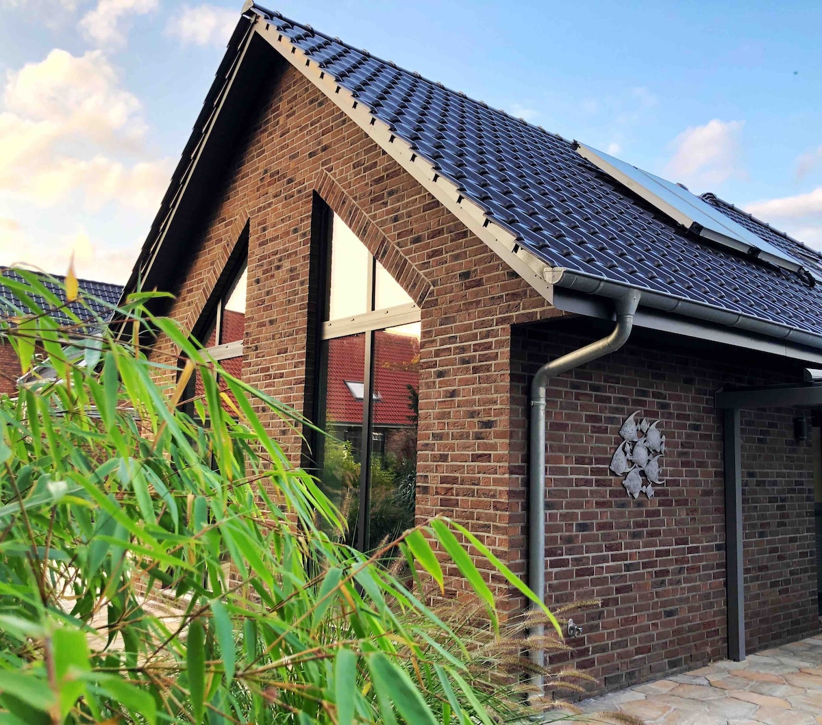 The exterior of the vacation home showcases a modern design with a sloped roof and large windows allowing natural light. The brick facade is complemented by lush greenery in the foreground, creating a serene and inviting atmosphere.