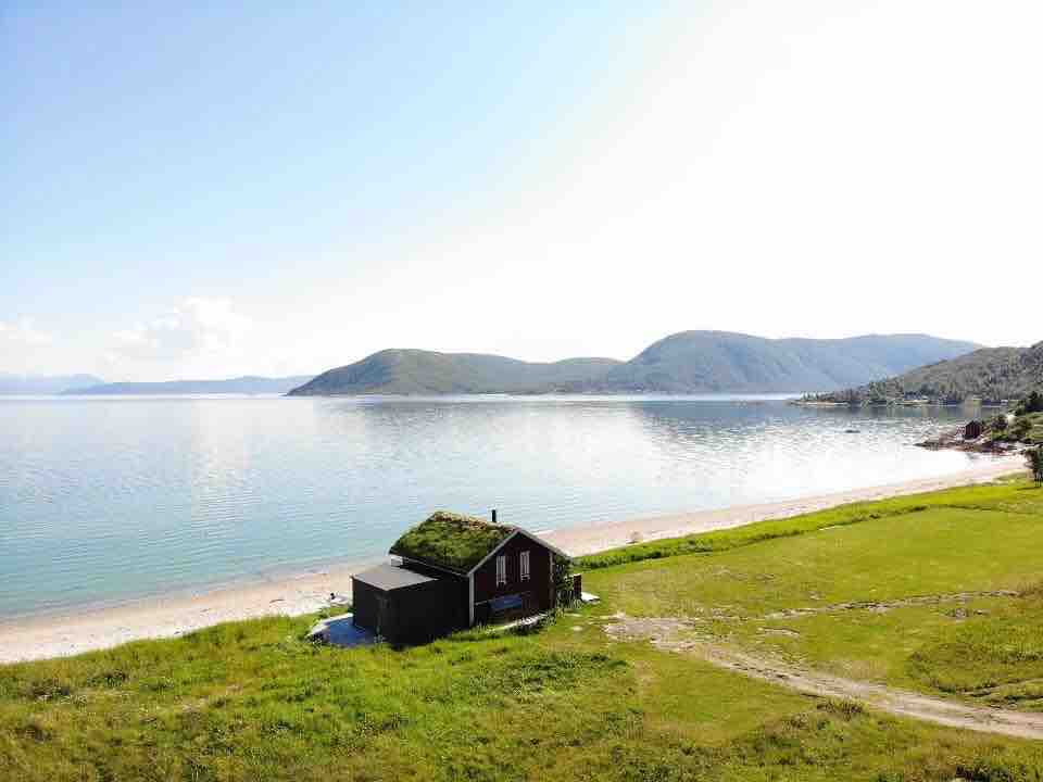 A quaint red cabin with a grass-covered roof is positioned near a calm body of water, surrounded by open green spaces. Gentle hills rise in the background under a clear blue sky, creating a serene coastal landscape.