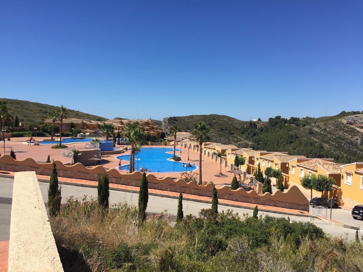 A spacious pool complex is depicted, featuring two large infinity pools surrounded by palm trees. Sun loungers are spread around the deck, with well-maintained gardens accentuating the area. The buildings in the background blend into the hillside, providing a scenic contrast against the clear blue sky.