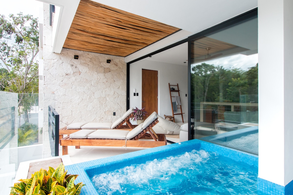 A private terrace features a plunge pool with jets, surrounded by textured stone walls and a wooden ceiling. A lounge chair is positioned beside the pool, inviting relaxation. Floor-to-ceiling glass doors provide a seamless view of nature while maintaining an open feel.