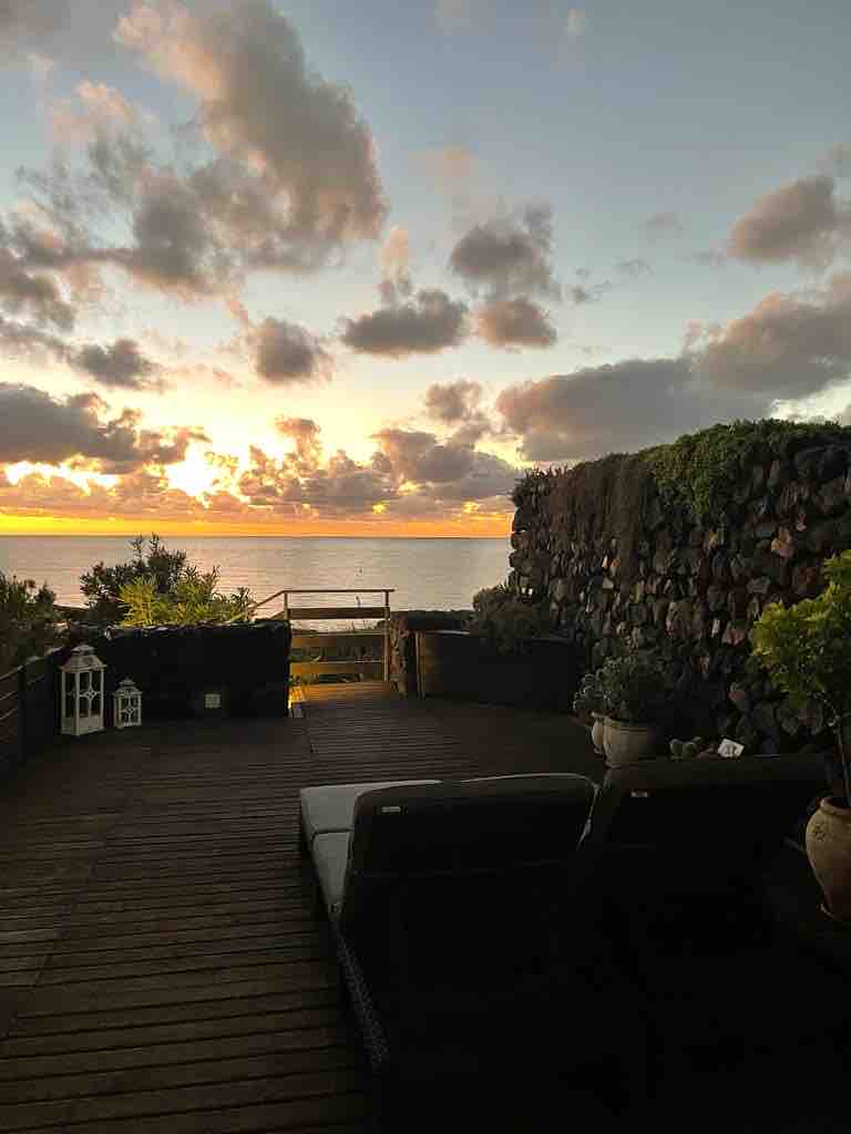 A terrace area features wooden flooring and plants along a stone wall. Two lounge chairs are positioned for sunset views over the sea, with the horizon illuminated by warm colors. Decorative lanterns add a touch of charm to the setting.
