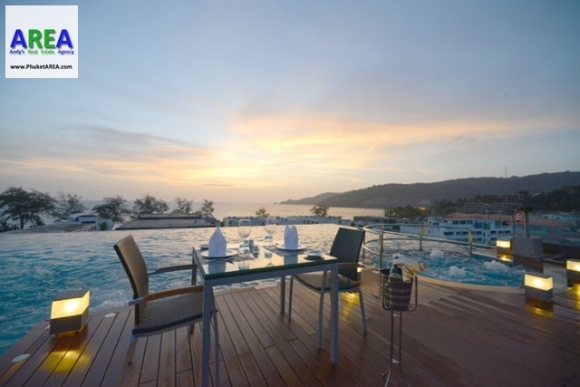 A dining area is set on a wooden deck beside an infinity pool, offering panoramic views of the ocean during sunset. A table is neatly arranged with utensils and wine glasses, while soft lighting enhances the serene atmosphere.
