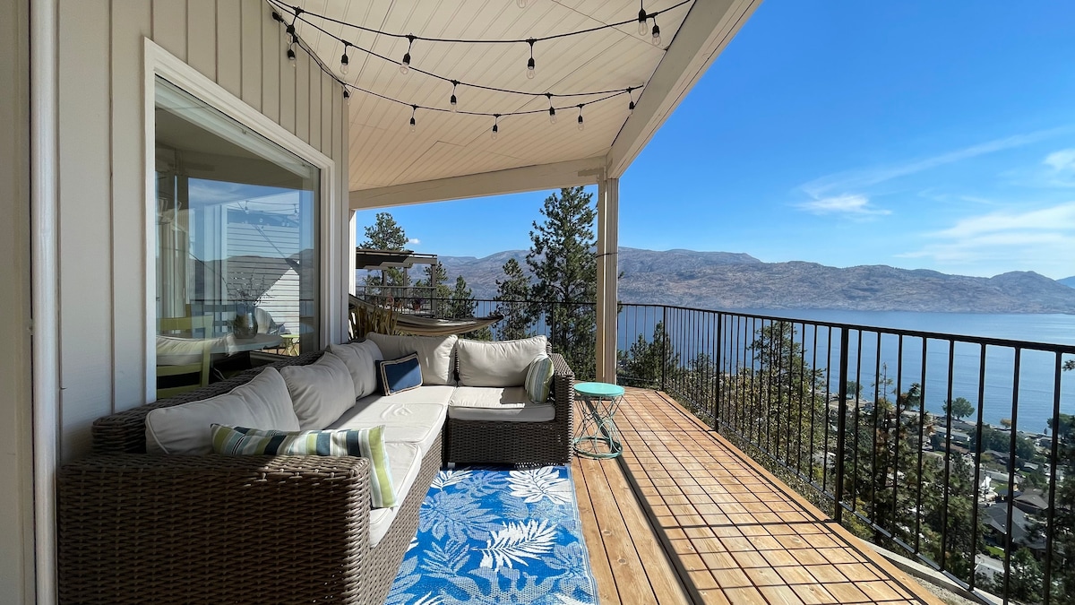A large covered deck features outdoor furniture arranged for relaxation, with a vibrant area rug underneath. Strings of lights hang overhead, providing ambient illumination. The unobstructed view of Okanagan Lake and surrounding mountains is prominently displayed, enhancing the outdoor living space.