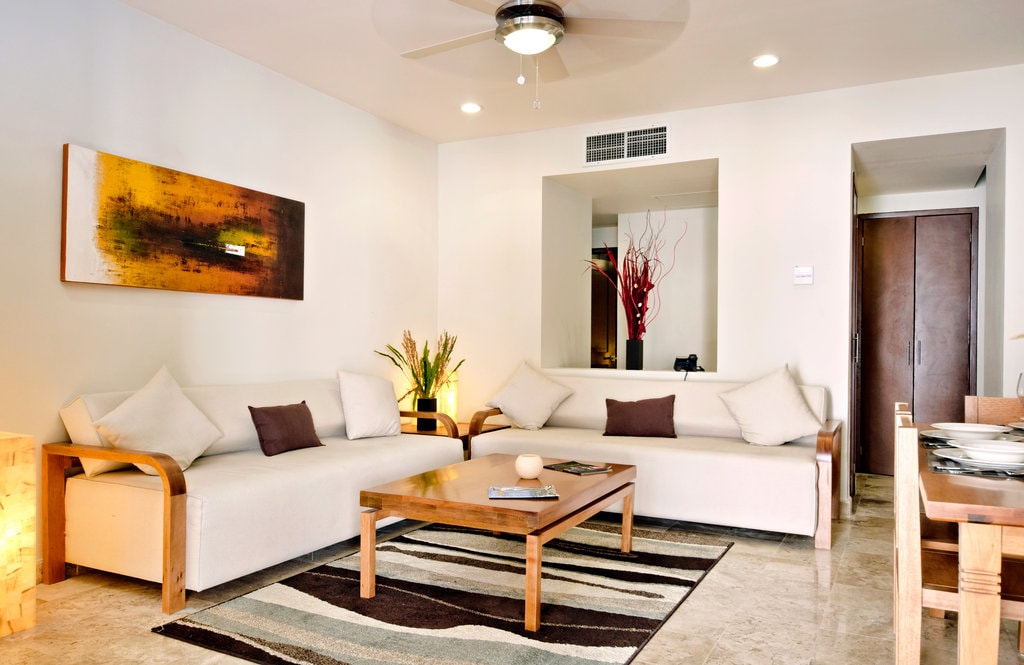 A bright living area features a comfortable seating arrangement with light-colored sofas complemented by decorative pillows. A wooden coffee table sits at the center on a striped rug, while natural light streams through nearby windows, and decorative plants add a touch of greenery.