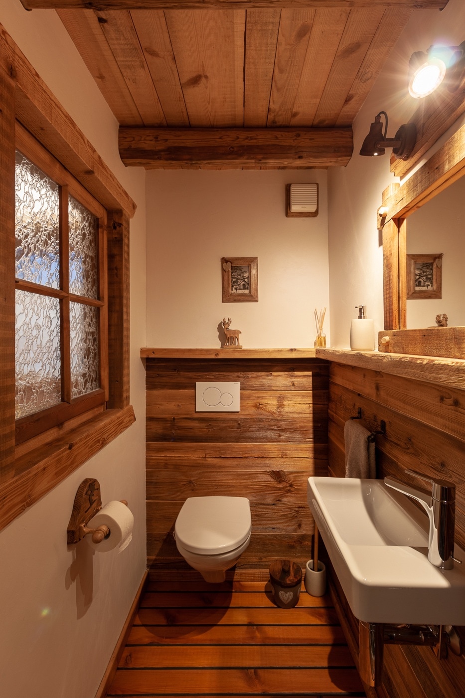 The bathroom features a compact layout with wooden walls and flooring. A wall-mounted sink is accompanied by a toilet, both positioned beneath warm lighting. Natural elements are incorporated, including a decorative wooden figure and an artistic window that provides a glimpse of the outside.