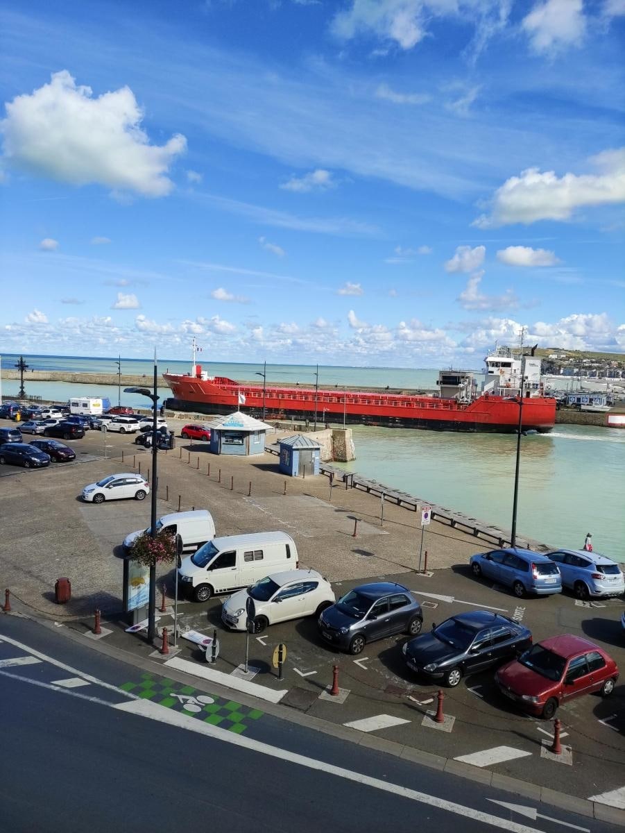 A view of the harbor from the apartment, showcasing a cargo ship docked alongside. Vehicles are parked along the waterfront, with a mix of cars and a van visible. Scenic clouds drift across a blue sky, enhancing the maritime atmosphere of the area.