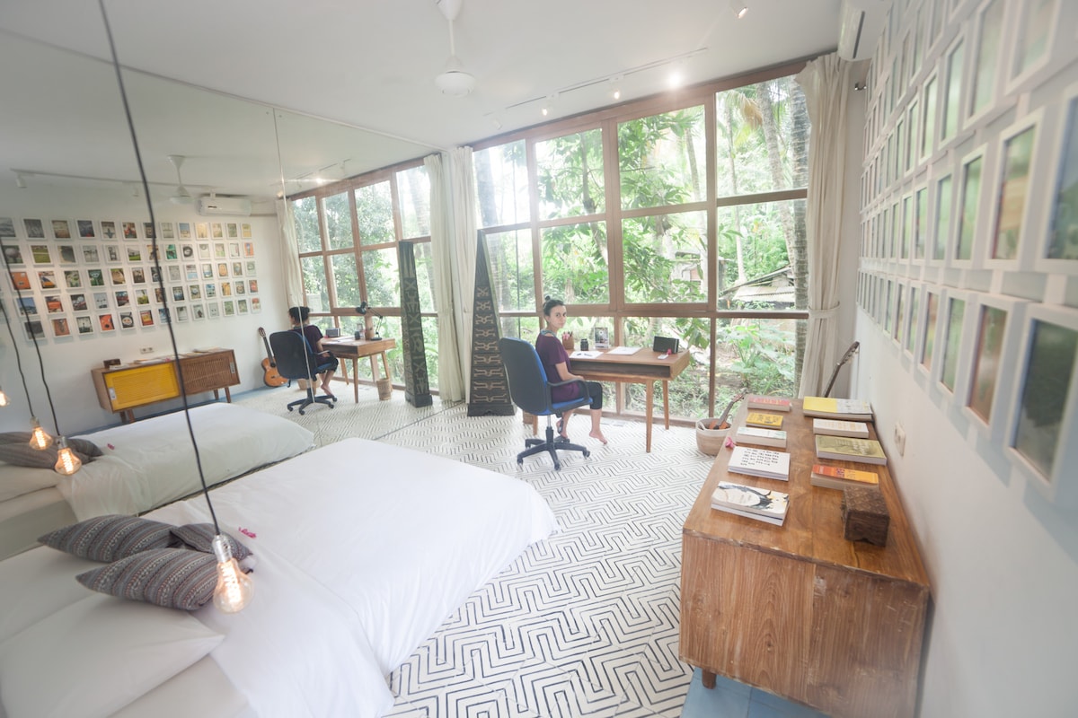 A spacious room is bathed in natural light from large windows, surrounded by lush greenery. A minimalist design features a clean, white bed with soft bedding. A teak desk sits adjacent to the window, providing a view of the outside. Artworks line the walls, adding a creative touch.