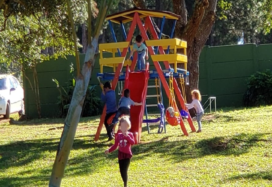 A colorful playground is set within a grassy area, featuring a slide and climbing structure. Children are engaged in play, with some sliding down the red slide while others climb. Green trees surround the space, providing a natural backdrop and shade.