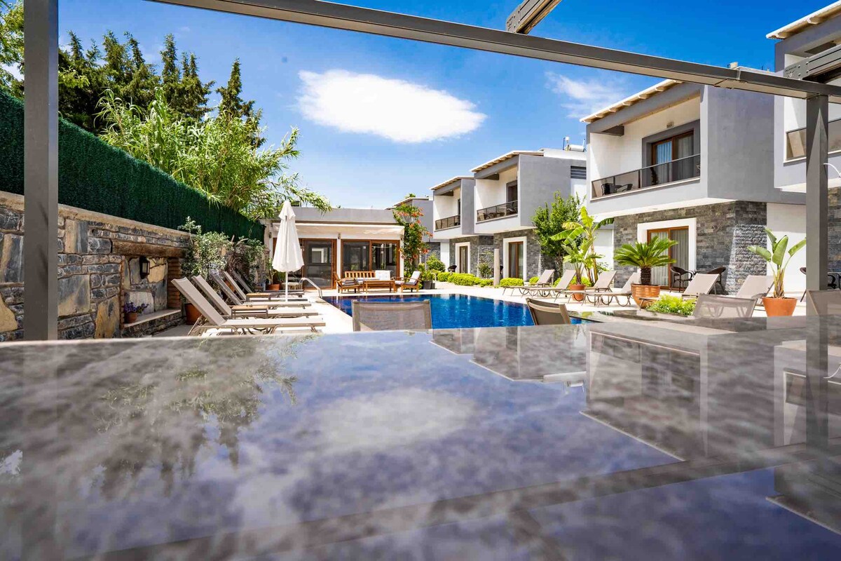 A serene outdoor area features a swimming pool surrounded by sun loungers and lush greenery. Two-story residences are visible in the background, framed by a clear blue sky. A covered area is also present, with shaded seating and potted plants enhancing the space.