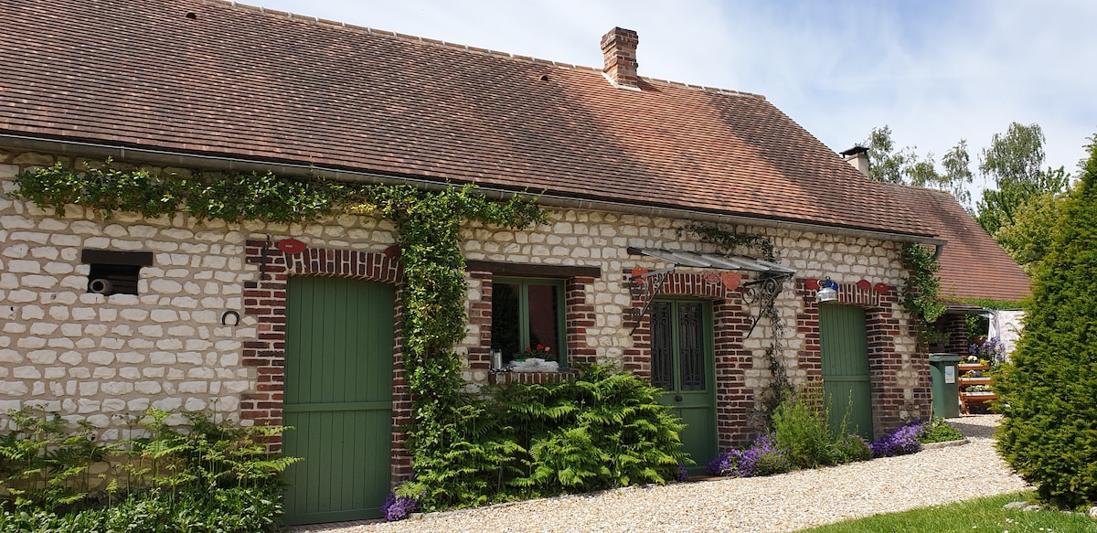 The renovated barn features a rustic façade with exposed brick and stone walls. Green doors are framed by climbing plants and purple flowers, creating an inviting entrance. A gravel pathway leads to the front, surrounded by well-maintained greenery.
