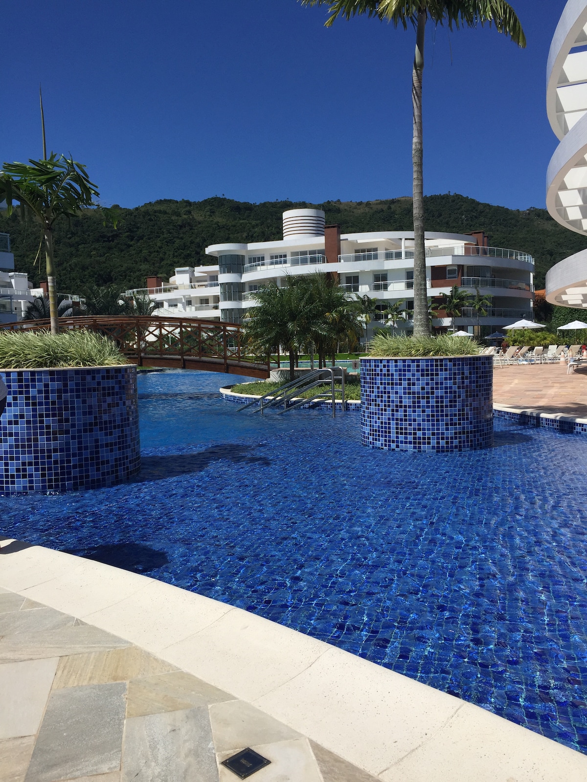 A resort-style swimming pool is surrounded by lush greenery, with blue mosaic tiles reflecting sunlight. A wooden bridge crosses over the water, leading to landscaped areas. The backdrop features modern buildings and palm trees, enhancing the tropical ambiance.