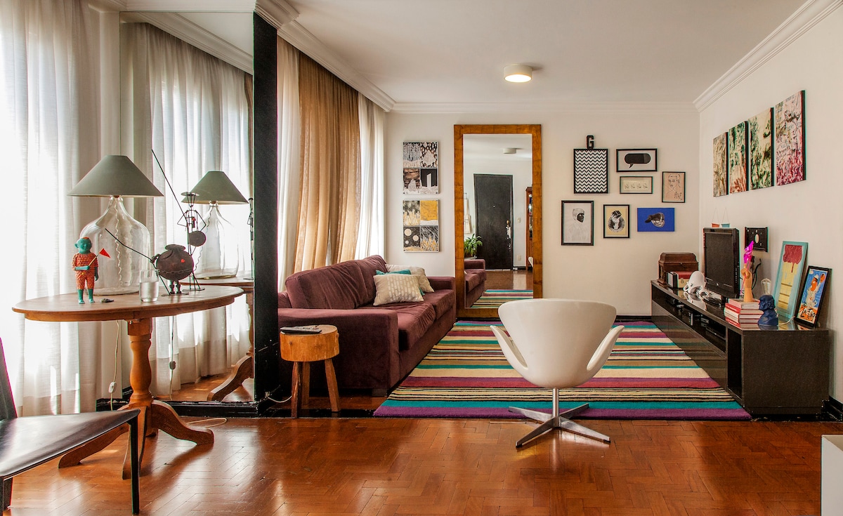 A spacious living area features natural light filtering through large windows draped with sheer curtains. A colorful striped rug anchors the space, which includes a modern white chair, a round wooden table, and a plush sofa, creating an inviting gathering spot.