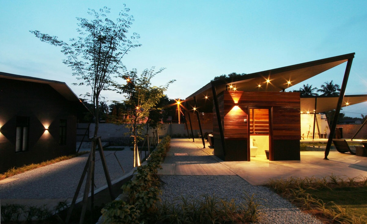 A modern garden villa is featured in an evening setting, illuminated by soft outdoor lighting. The villa's sleek design includes wooden accents and angular rooflines. Lush greenery and gravel pathways surround the structure, enhancing the tranquil ambiance of the private retreat.