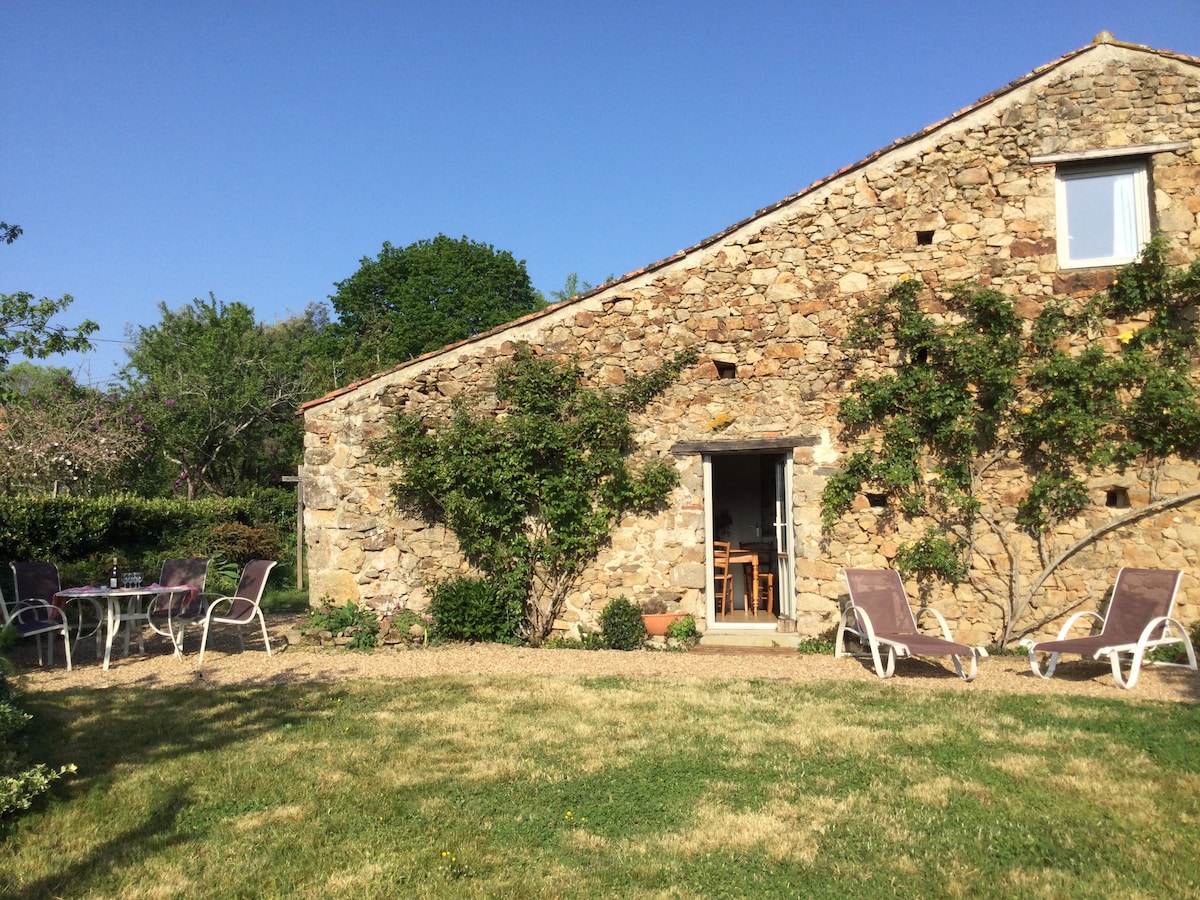 A stone gîte features a natural exterior with scattered shrubs and trees surrounding the property. A cozy outdoor area includes sun loungers and a dining set. A single window and double doors lead into the gîte, inviting a view of the interior.