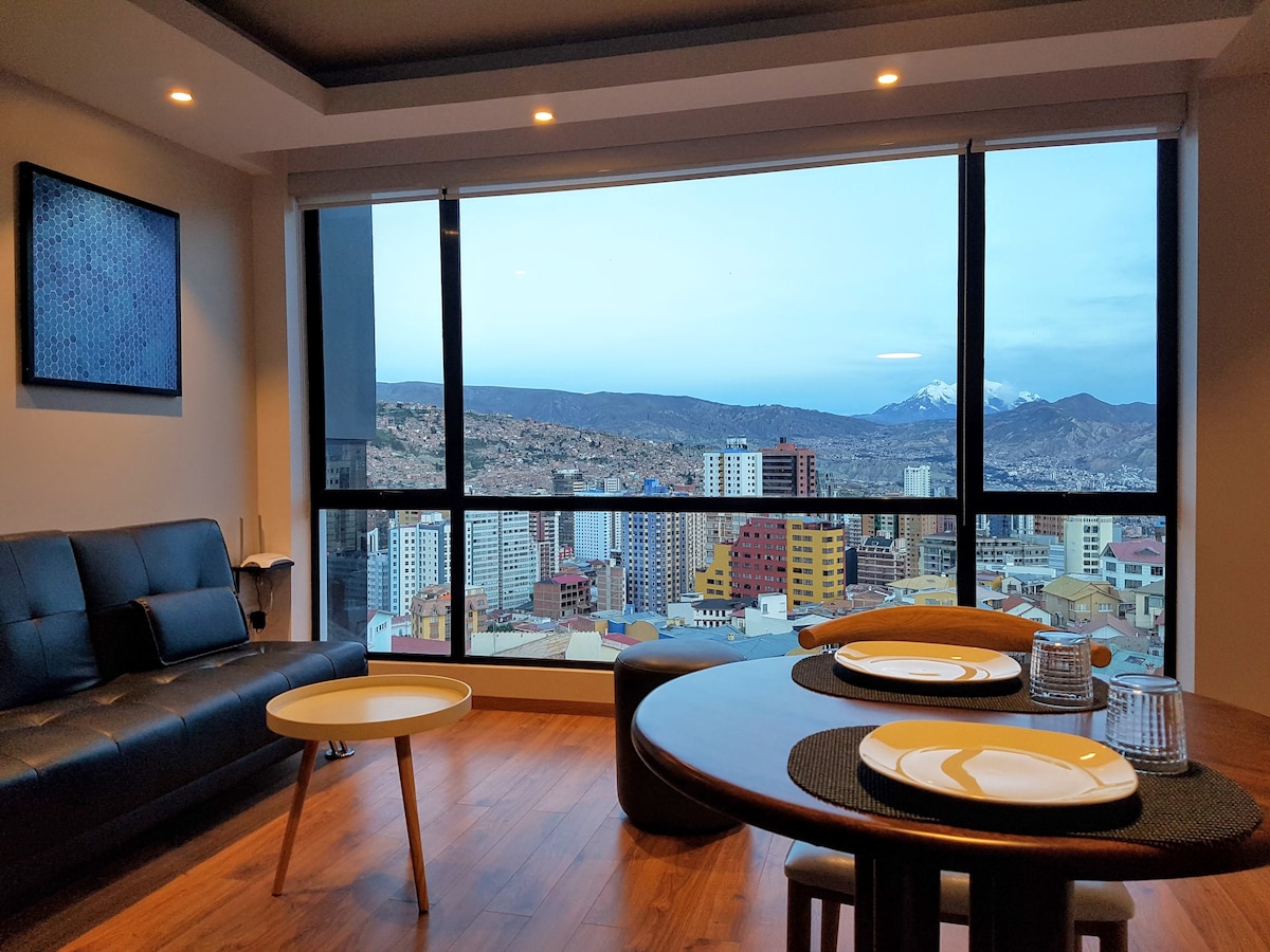 A modern living space features a large window offering panoramic views of the city. A sleek black sofa and round dining table are arranged to create a comfortable setting, complete with place settings for two. Warm wood flooring adds a touch of elegance.