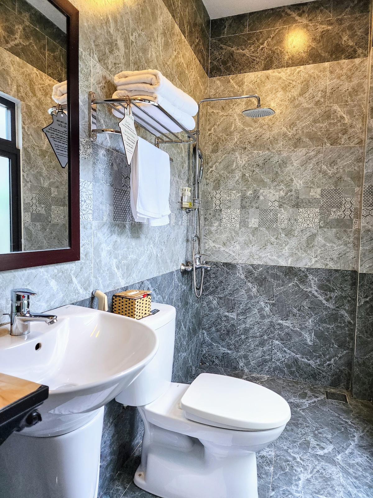 A modern bathroom features elegant gray marble walls and floor. A white porcelain toilet sits beside a sleek sink. A shower with a glass enclosure is equipped with adjustable fixtures. Fresh towels are neatly arranged on a rack above the sink.