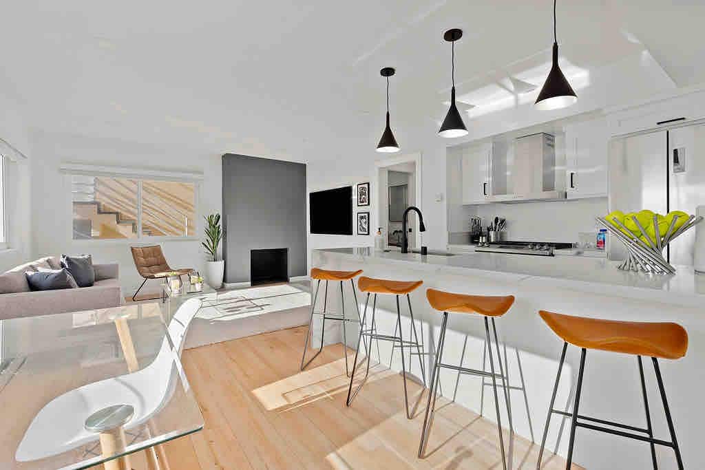 The modern kitchen features sleek white cabinetry and a central island with seating for four. Pendant lights hang above, highlighting the space. A gas fireplace adds warmth to the inviting area, while large windows offer natural light and a view of the nearby living area.