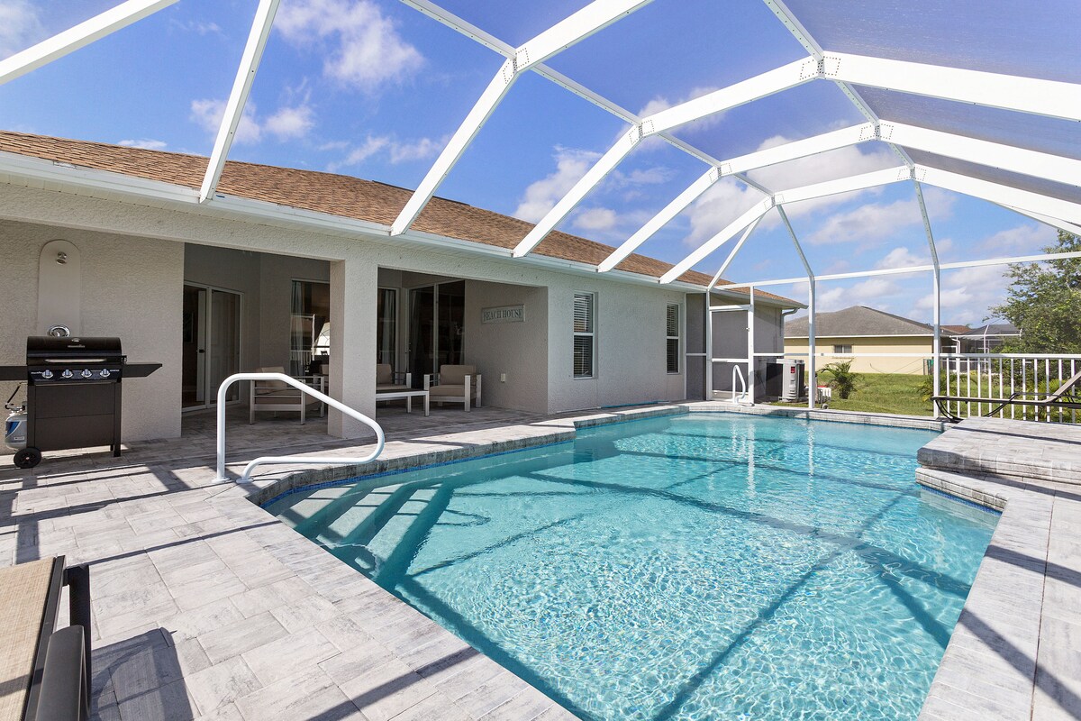 The outdoor area features a heated swimming pool surrounded by a screened lanai, with modern tile flooring. A grill is positioned nearby, and ample seating is available for relaxation. The pool area is bathed in natural light, providing a serene setting for leisure activities.