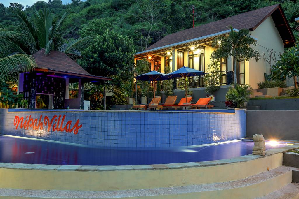 A spacious villa is set against a lush hillside, with an inviting infinity pool in the foreground. Sun loungers are positioned beside the pool, and umbrellas provide shade. The villa features large windows, blending harmoniously with the surrounding greenery.