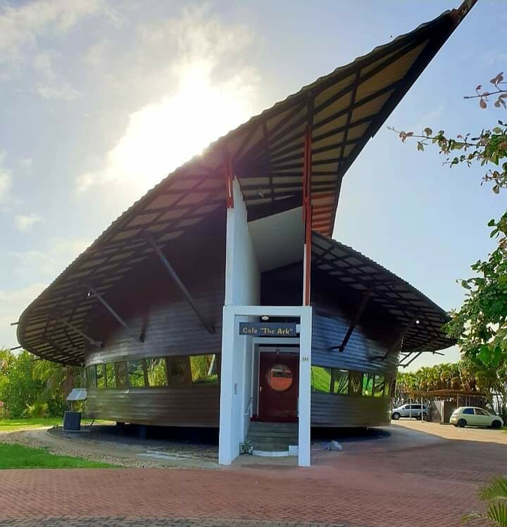 The exterior of De Ark restaurant features a unique, curved structure designed to resemble Noah's Ark. Made of tropical wood, it is set under a slanted roof that provides ample natural light. Large windows line the building, enhancing the connection to the surrounding environment.