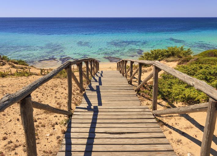 A wooden pathway leads down towards a serene beach, flanked by natural coastal vegetation. The clear blue water extends into the horizon, contrasting with the sandy shore. Soft waves gently lap at the water's edge, creating a peaceful seaside atmosphere.