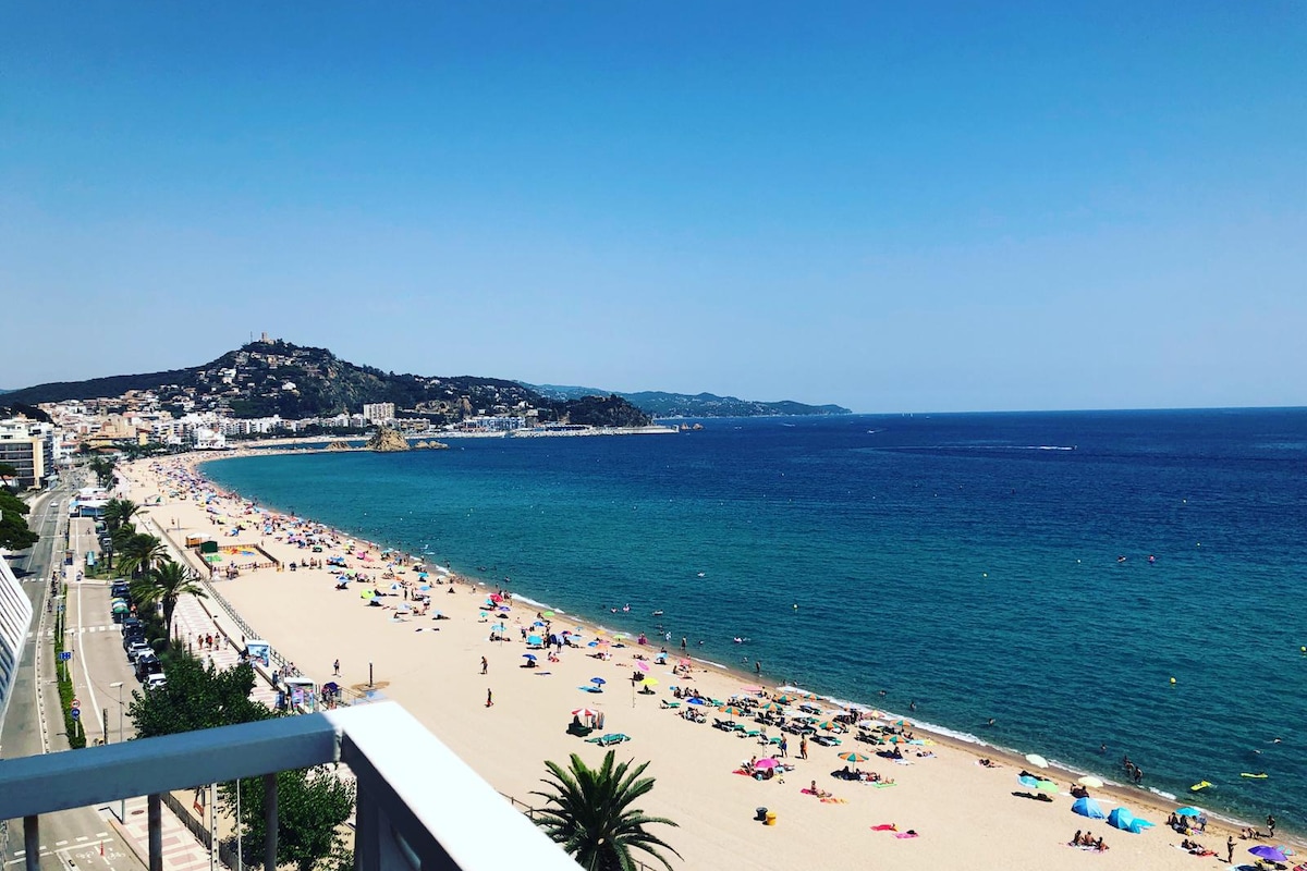 A panoramic view of the Mediterranean coastline, showcasing a sandy beach lined with colorful umbrellas and sunbathers. The clear blue water gently laps at the shore, while a distant hillside features a charming town and greenery, creating a serene seaside atmosphere.