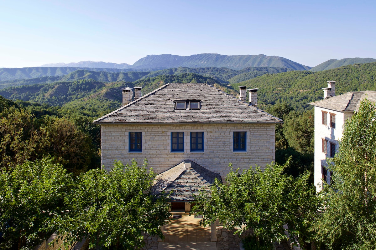 The exterior of a large stone house is seen, surrounded by lush greenery and trees. A scenic view of the mountains stretches in the background, showcasing rolling hills under a clear blue sky.