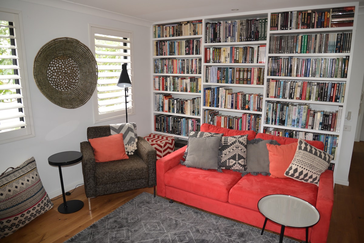 A private lounge area features a red couch adorned with patterned cushions, complemented by a cozy armchair. A round side table stands nearby, while a collection of books lines the shelves on the adjacent wall, illuminated by natural light through partially open shutters.