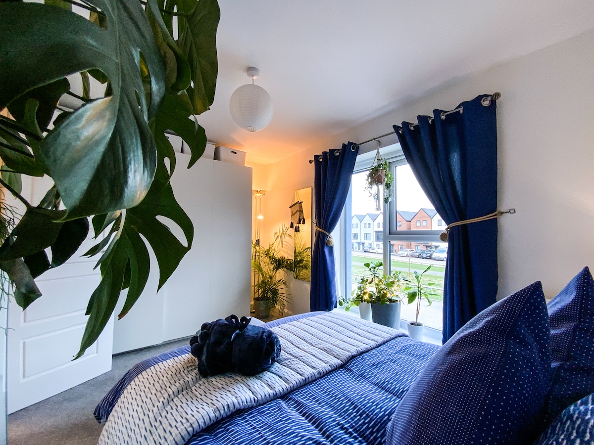 A cozy bedroom features a spacious bed layered with textured bedding. Large windows with blue curtains allow natural light to enter, while decorative plants add a touch of greenery. A white wardrobe stands against the wall, complementing the room's calming atmosphere.