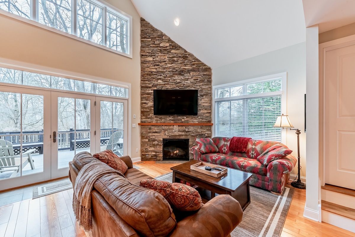 A spacious living area is showcased, featuring a stone accent wall with a television mounted above a fireplace. Comfortable seating is arranged around a wooden coffee table, with large windows allowing natural light to fill the space. A door leads to an outdoor deck.