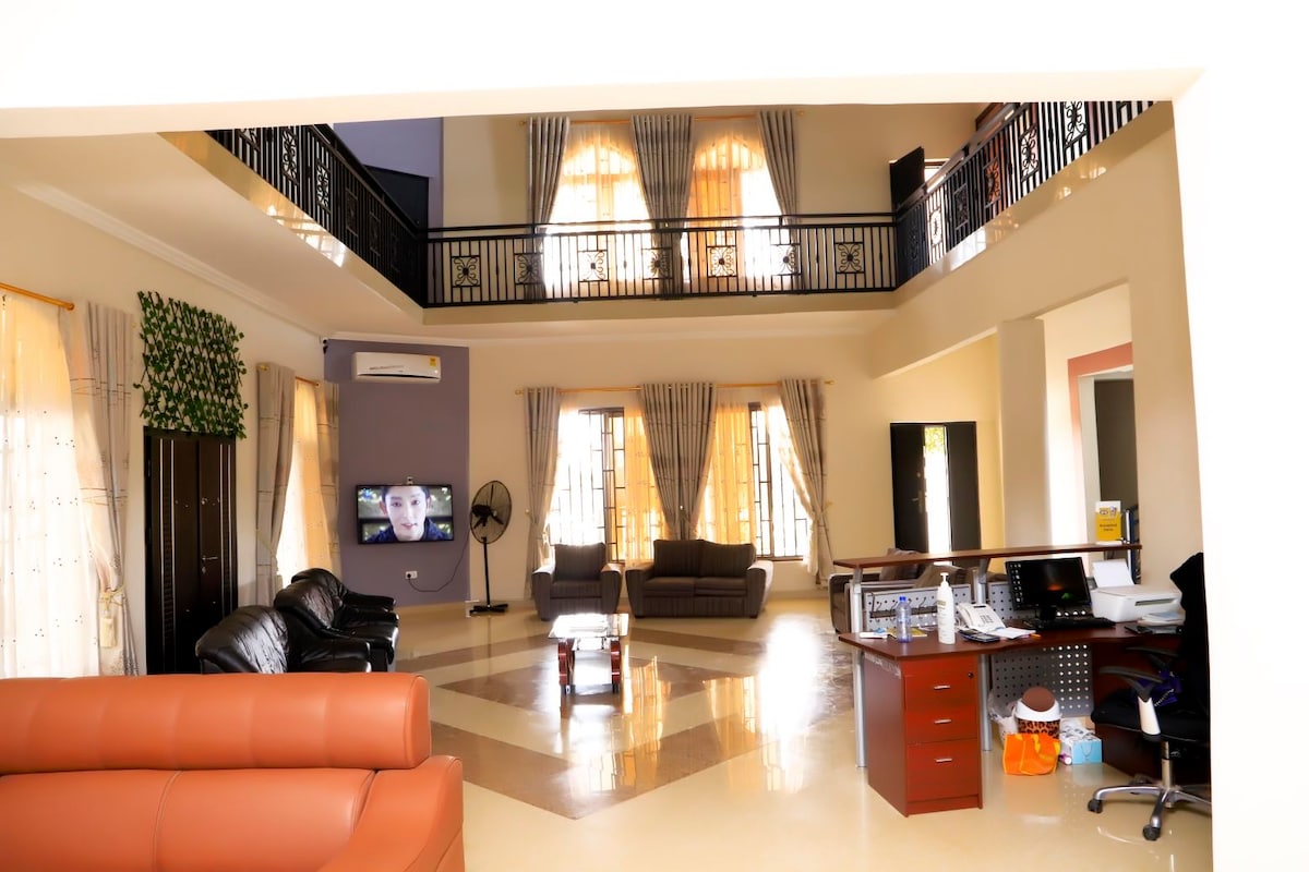 The spacious living area features high ceilings and an open floor plan, with large windows allowing natural light to fill the room. Comfortable seating options include a leather sofa. A balcony overlooks the space, enhancing its airy ambiance.