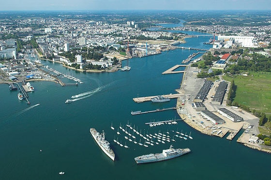 An aerial view showcases the Lorient harbor, with various boats anchored in the water. The shoreline features docks and a mix of industrial and commercial buildings. Lush green landscapes and urban areas can be seen in the background, framed by a clear blue sky.