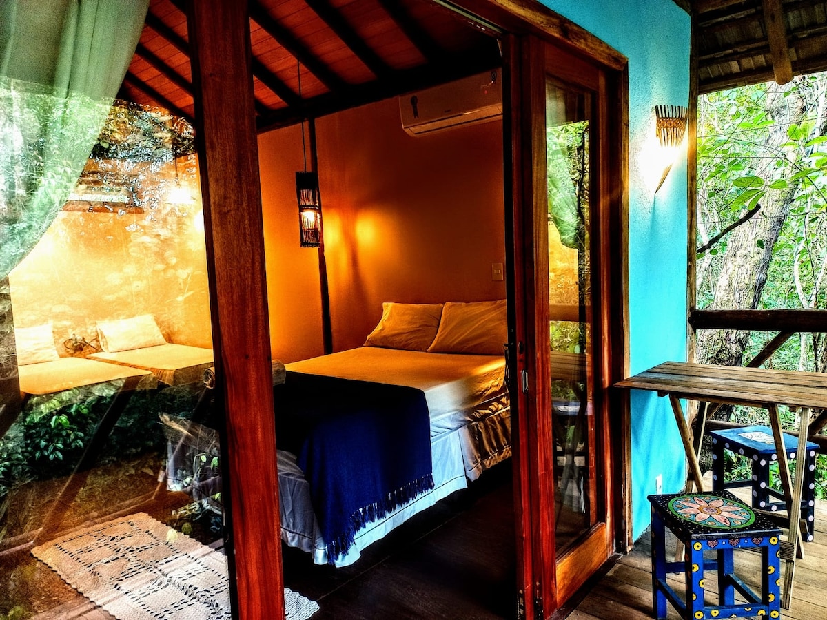 A spacious bedroom is surrounded by large glass doors that open to a lush outdoor view. The room features a comfortable bed with soft linens and a small round table with two chairs, adding to the inviting atmosphere. Natural light filters through the curtains.