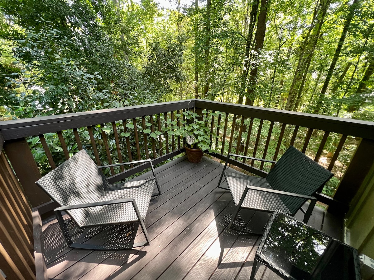 A private deck is surrounded by lush greenery, featuring two chairs positioned beside a small table. A potted plant adds a natural element, creating a peaceful space for relaxation in the wooded setting.