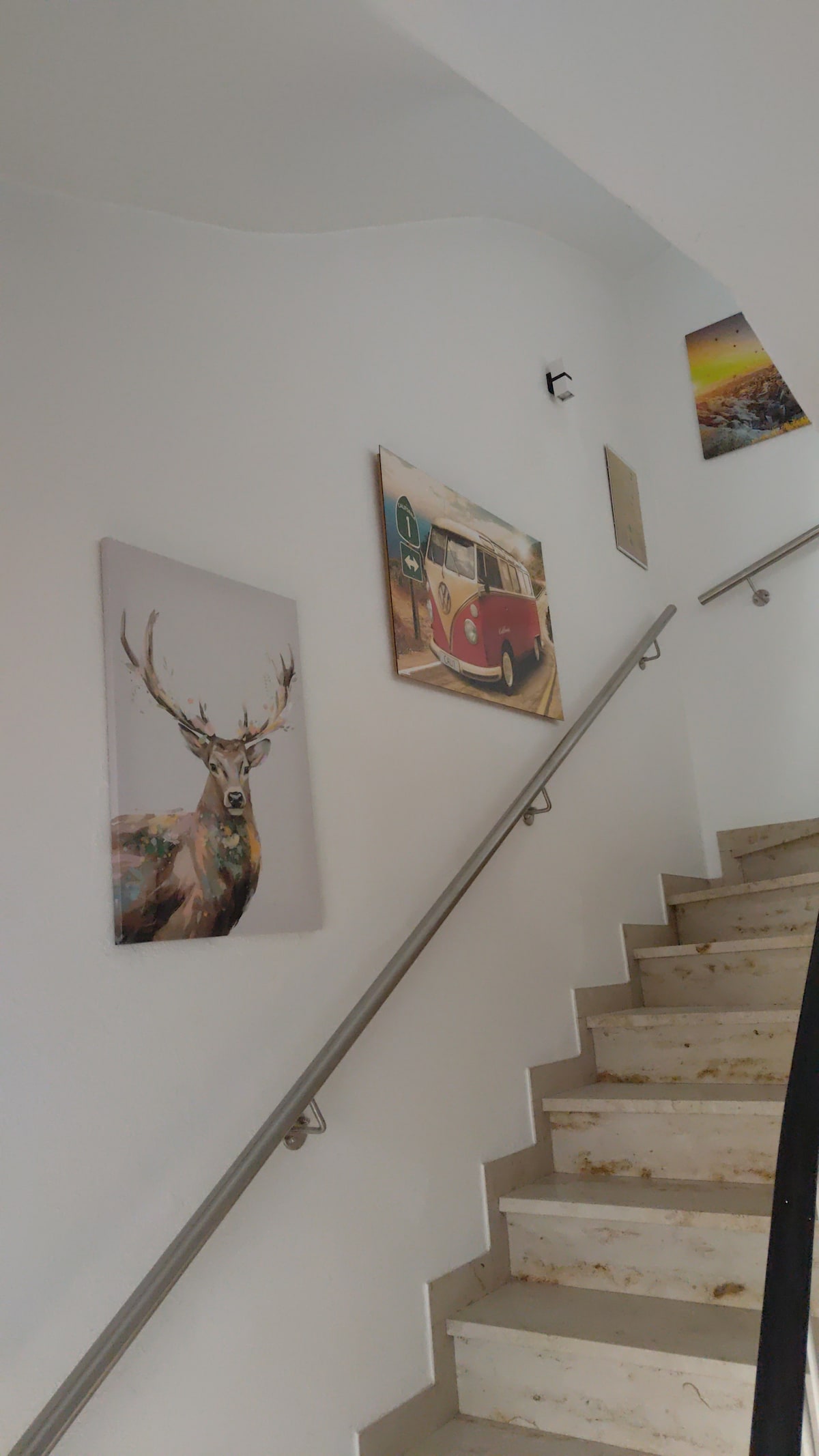 A light-filled stairway features a smooth handrail and marble steps. On the white walls, art pieces are displayed, including an image of a deer and an iconic vintage car, contributing to a welcoming ambiance.