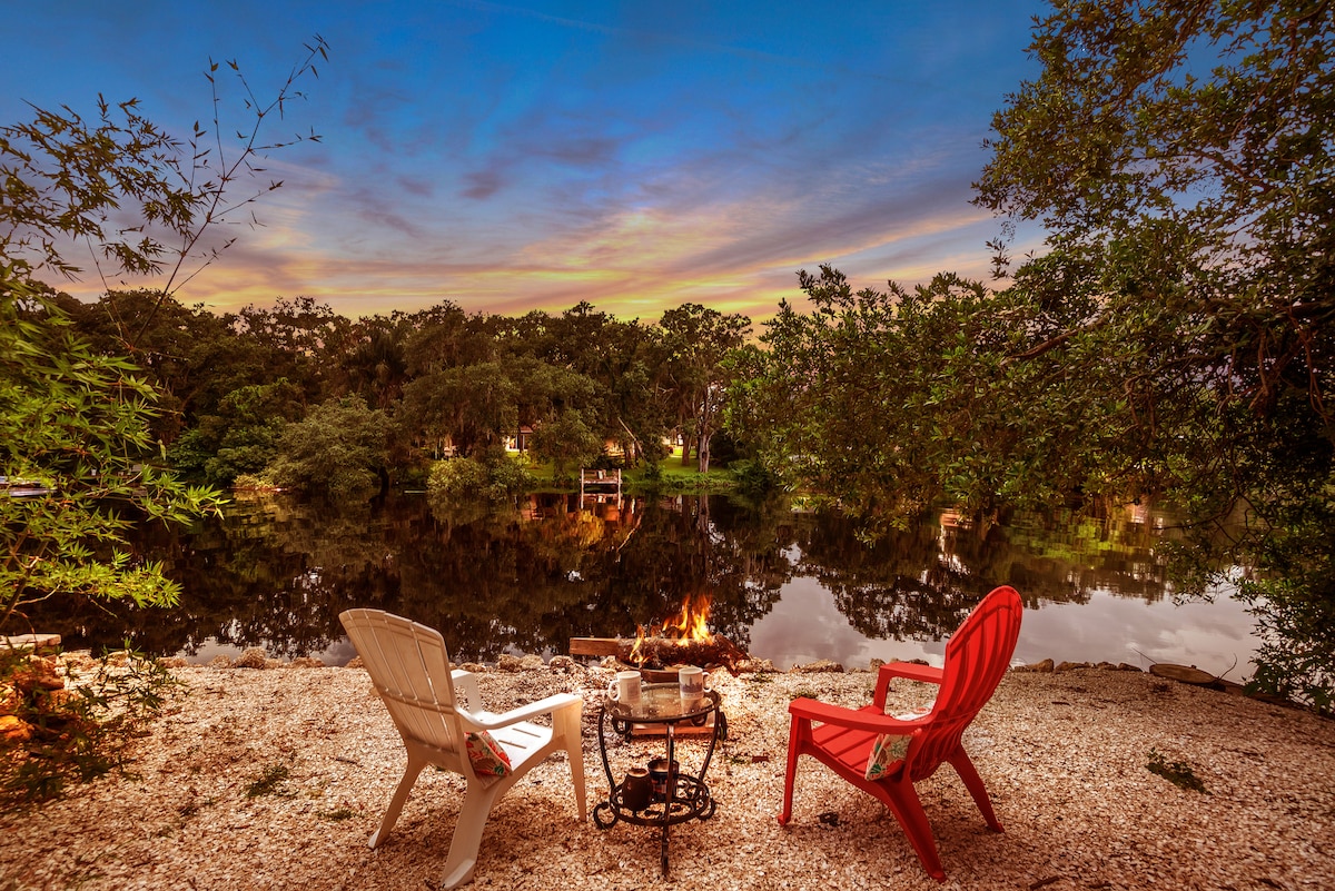 Two Adirondack chairs, one white and one red, are positioned on a pebbled area near a calm water body. A small fire pit is set between the chairs, surrounded by lush greenery and reflecting trees in the serene water, creating a peaceful outdoor setting.