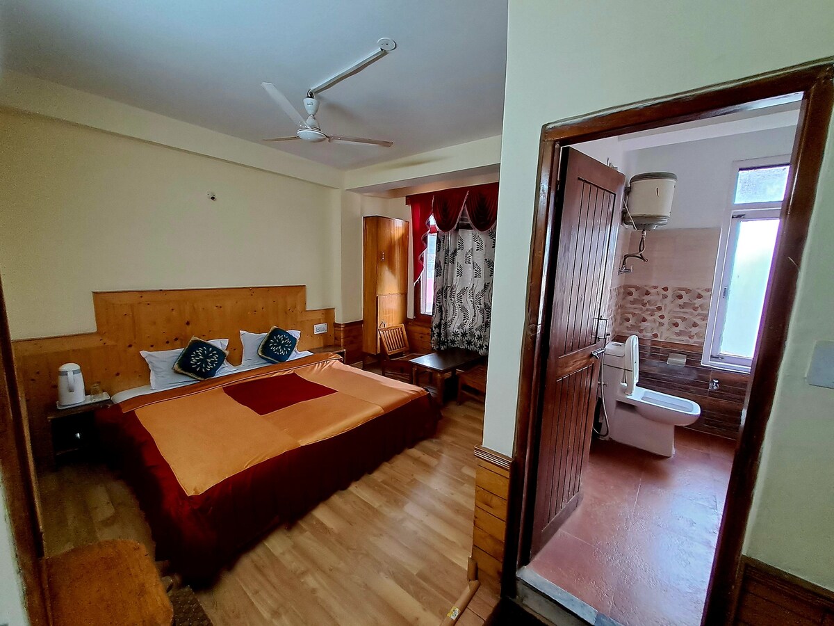 A room is presented featuring a spacious double bed with an orange bedspread. A small wooden table with two chairs is situated in the corner. A bathroom with modern amenities is accessible from the room. Natural light streams through the window covered by sheer curtains.