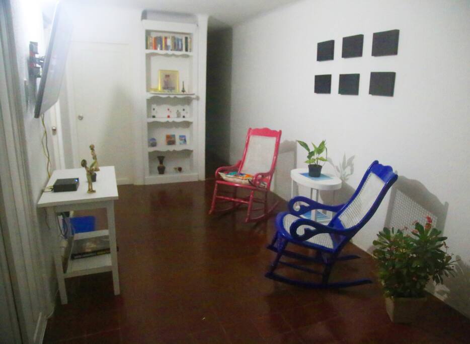 Living room equipped with a plasma TV, a desk, two tables, plants, chairs and a mantel.