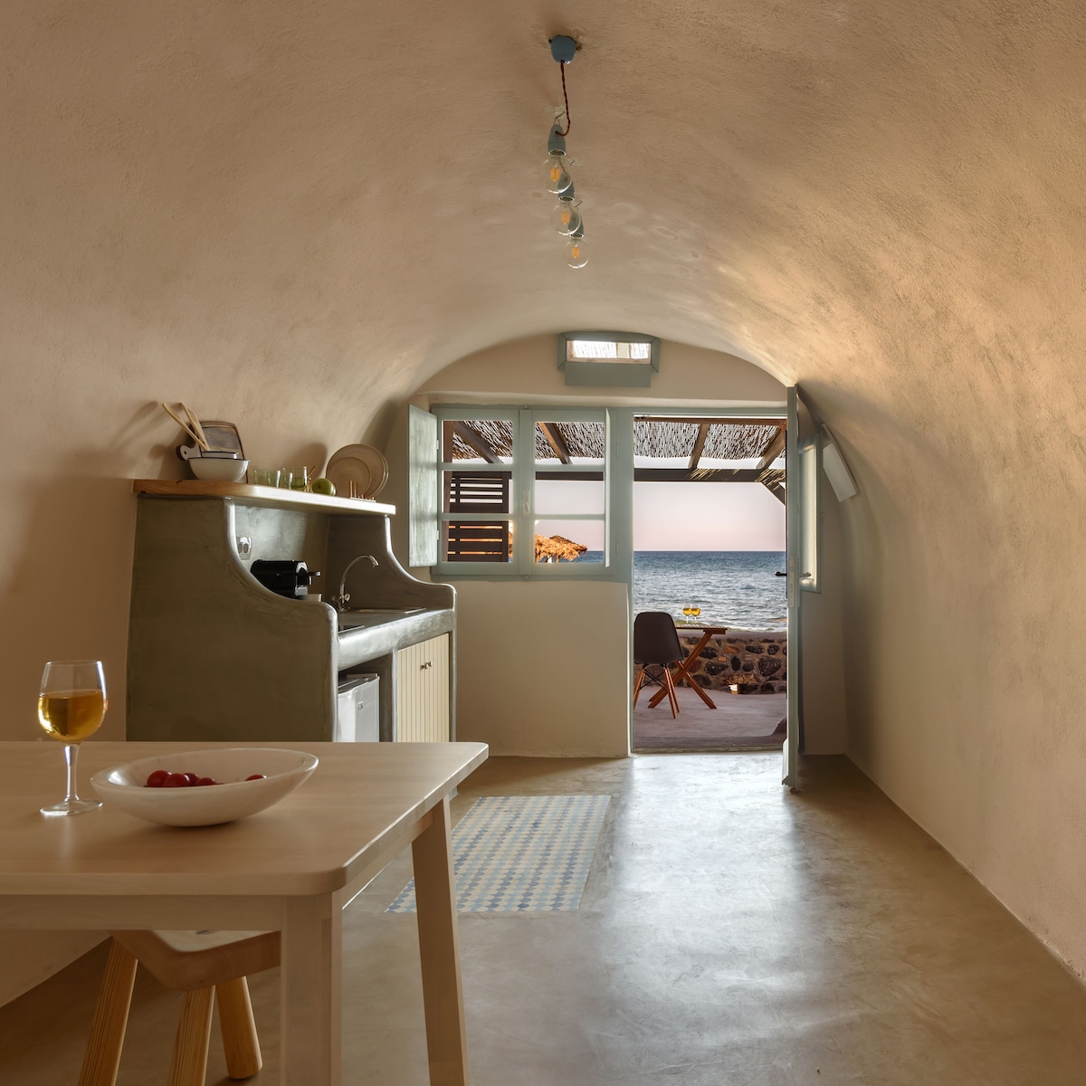 A charming kitchen area features a simple wooden table with a fruit bowl and a glass of wine. A green countertop and exposed light fixtures contribute to the cozy design. A doorway leads to a patio with ocean views, framed by open shutters.