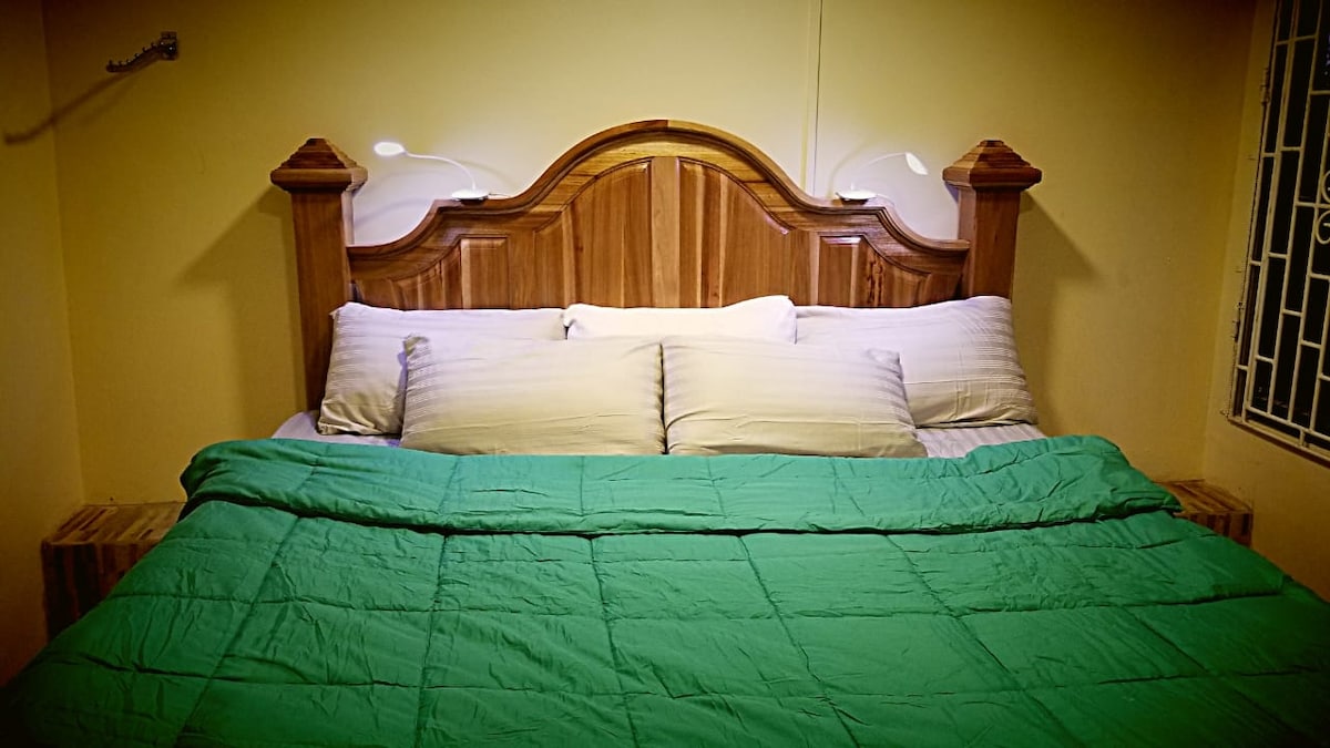 A spacious bed is centered against a softly lit wall, featuring a wooden headboard with side lamps for nighttime reading. Light and dark pillows are arranged neatly, with a vibrant green quilt layered over the bed, inviting a cozy and restful atmosphere.