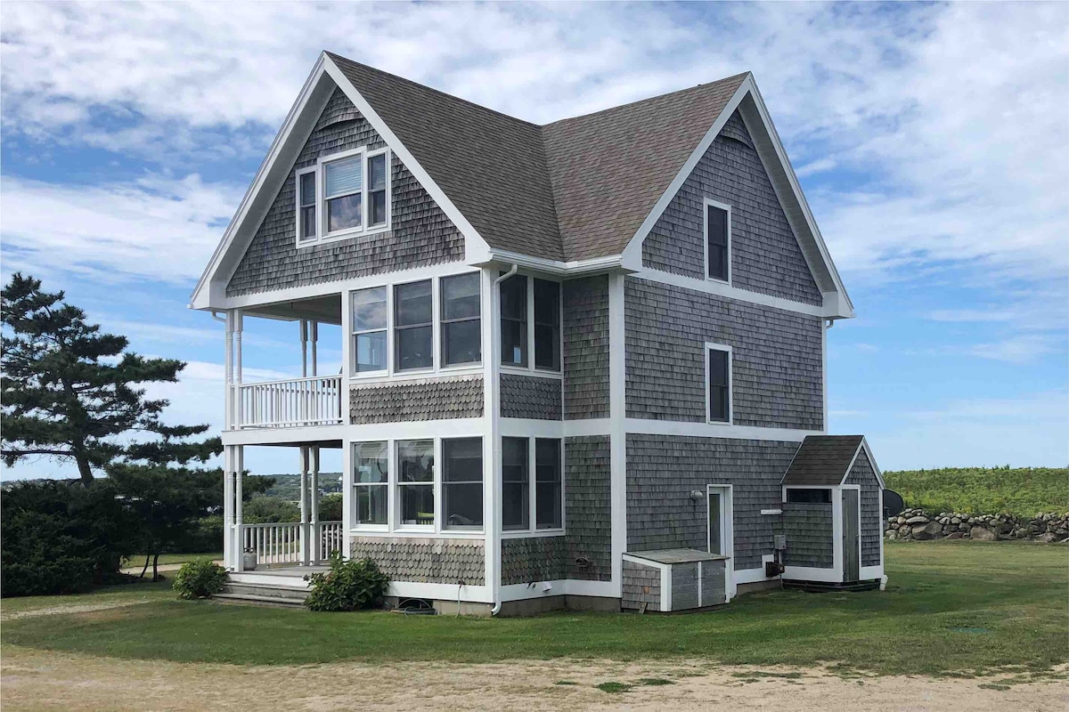 A three-story home with a shingle exterior is featured, showcasing multiple large windows that allow natural light. A porch extends across the first and second floors, and a small storage shed is visible to the right. Lush greenery surrounds the property.