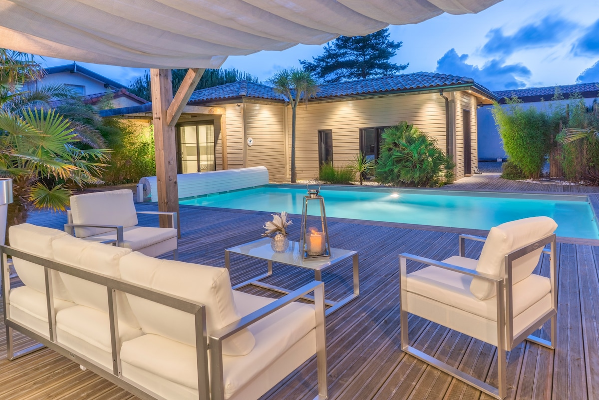 An outdoor seating area features modern white armchairs arranged around a glass table, shaded by a retractable awning. A tranquil pool is visible in the background, surrounded by tropical plants and wooden decking, providing a serene atmosphere for relaxation.