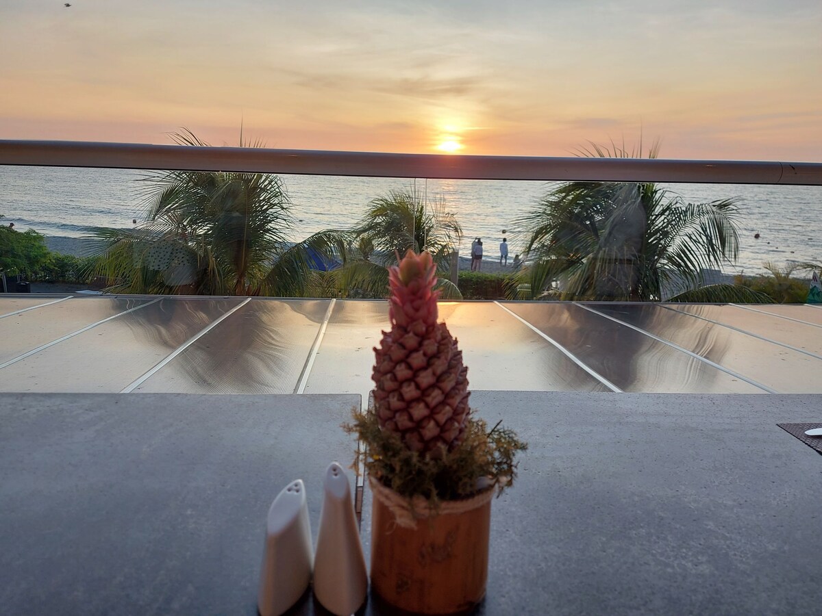 A small decorative pinecone is placed in a pot on a table, set against a backdrop of a sunset over the ocean. The scene includes palm trees and gentle waves, with figures walking along the beach in the distance.