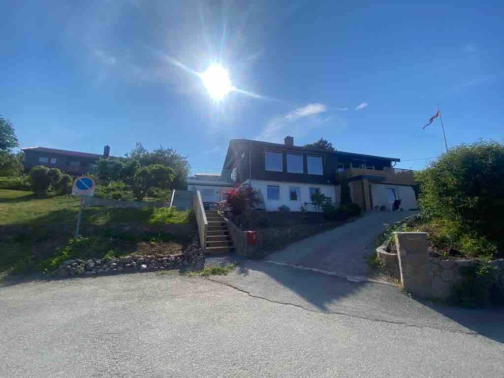 A spacious family-friendly home is set against a clear blue sky, featuring a welcoming entrance with steps leading up from the driveway. Lush greenery surrounds the property, while sunlight casts a warm glow over the exterior.