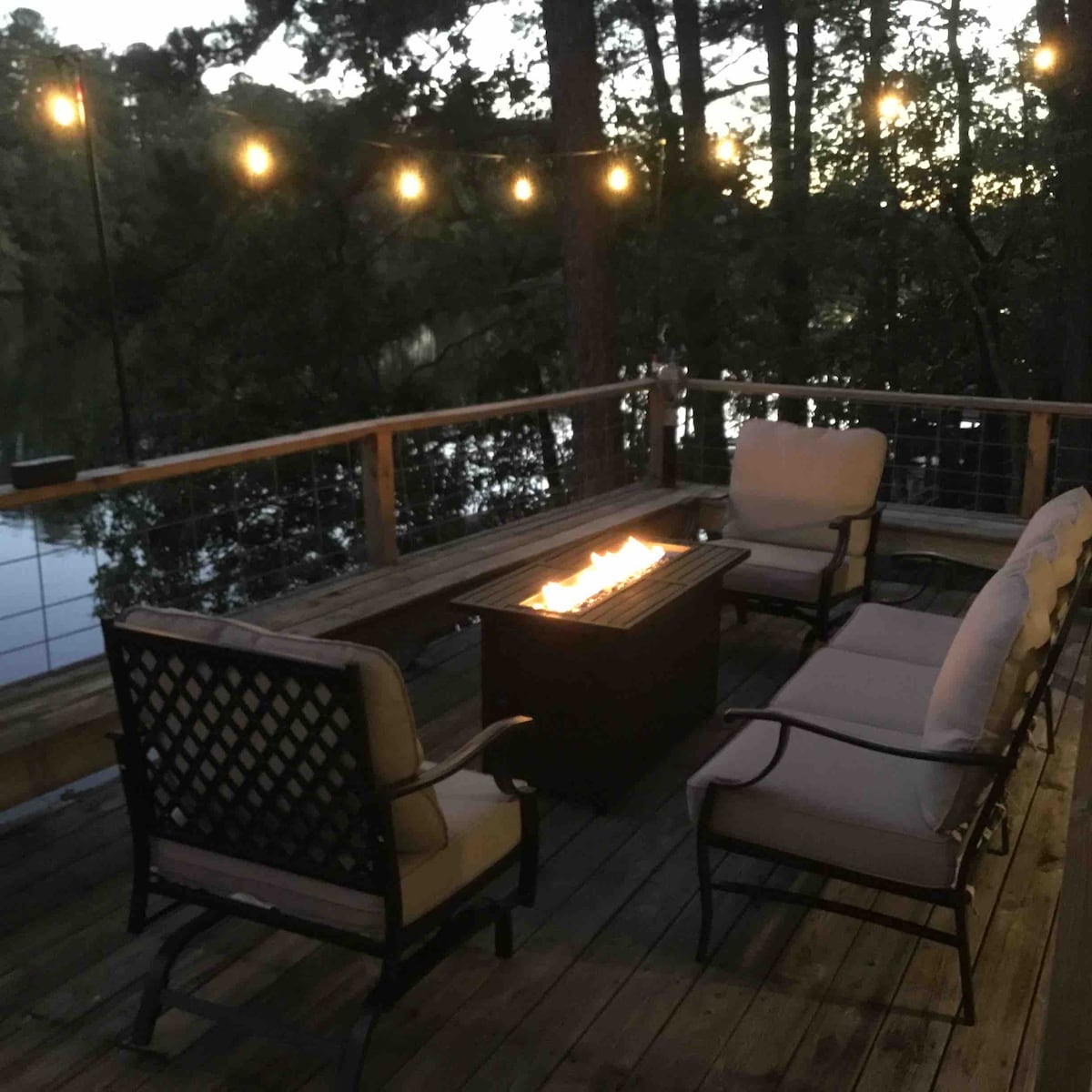 A cozy outdoor seating area is featured on a wooden deck surrounded by trees. A fire pit is centered among comfortable chairs, creating a soothing ambiance. String lights are hung above, illuminating the space gently as dusk sets in, with a view of the lake nearby.
