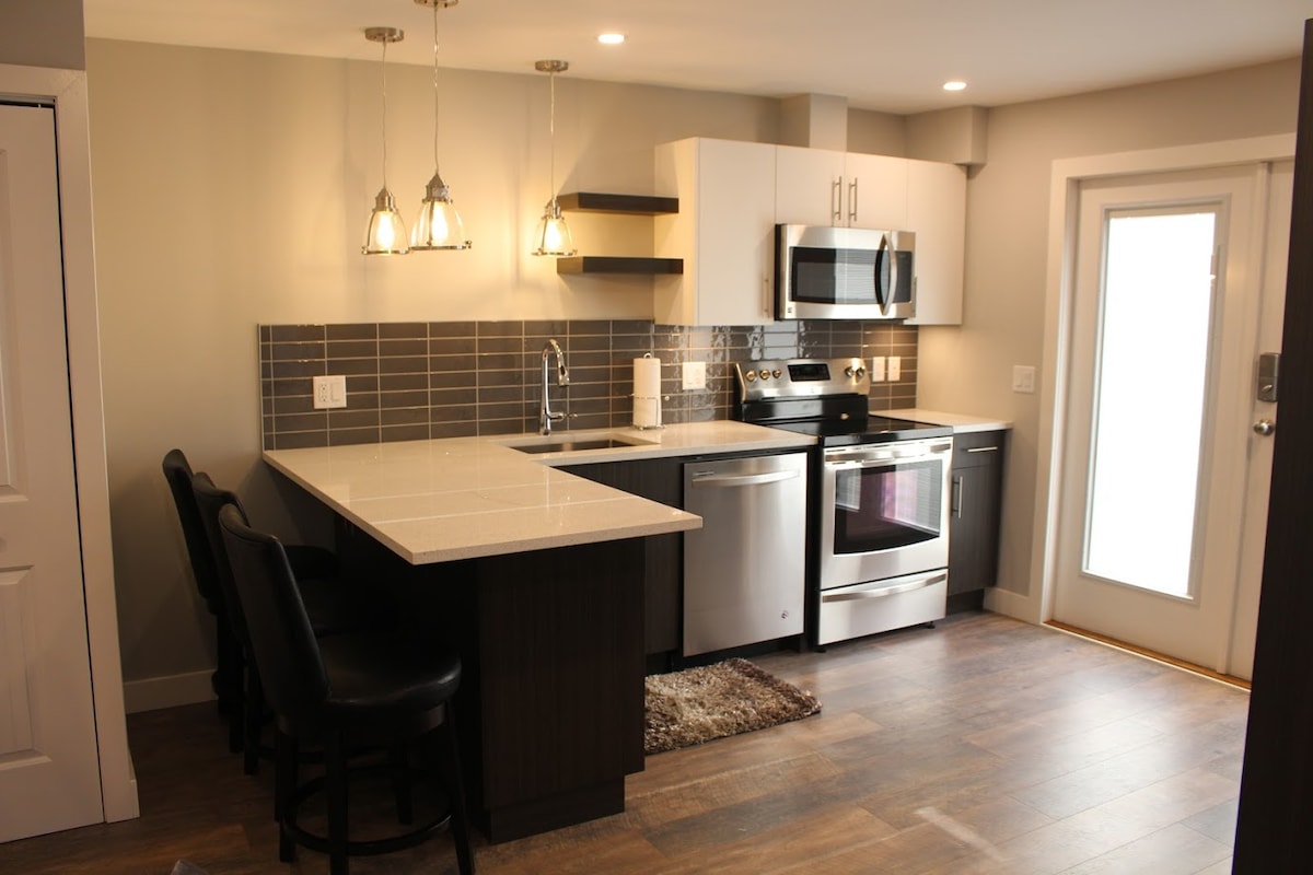 The kitchen features modern stainless steel appliances, including a refrigerator, oven, and microwave. A spacious island with bar seating for four is provided. Pendant lights hang above, illuminating the countertops and stylish backsplash, while a door leads to an outdoor area.
