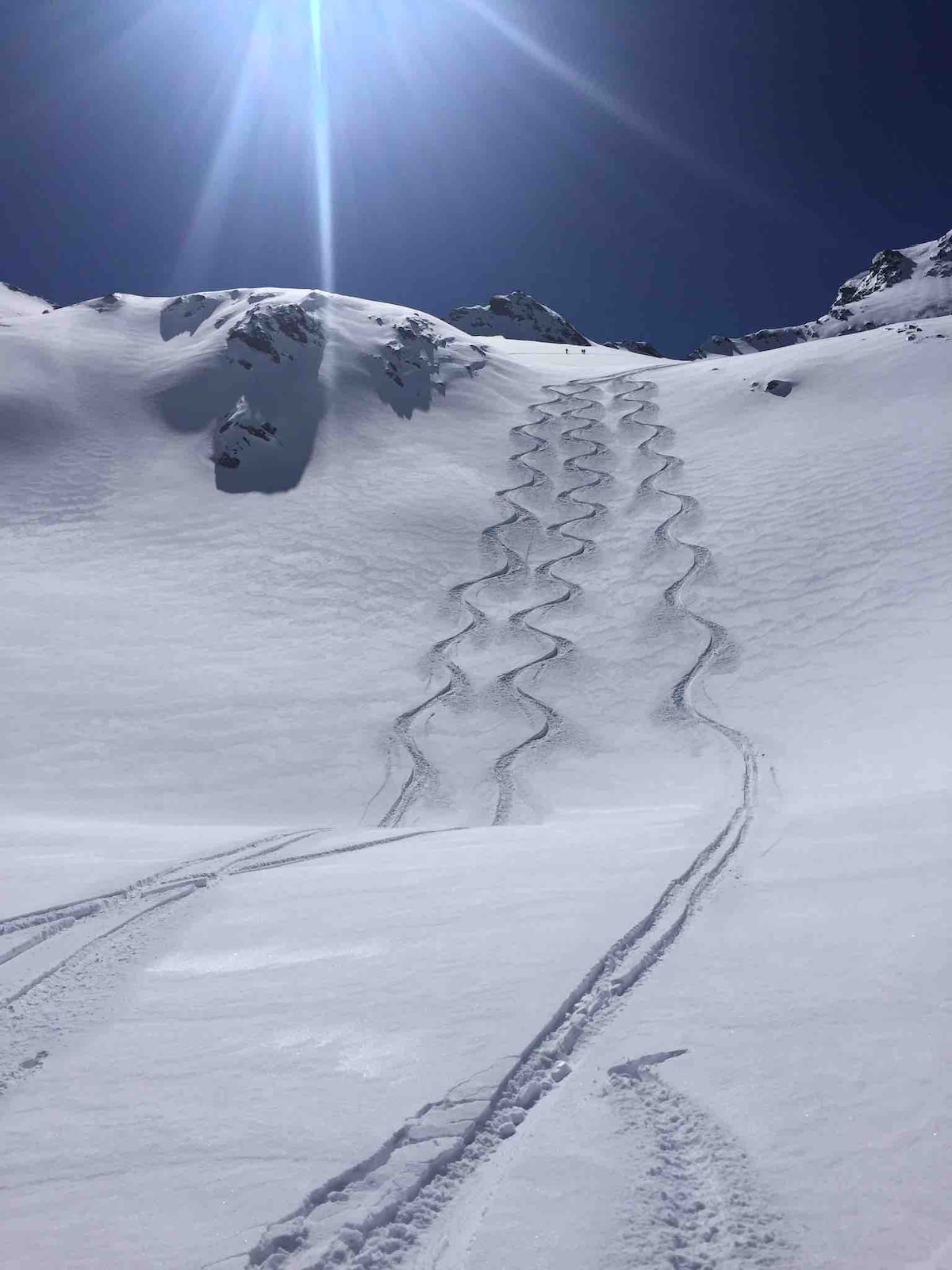 A snowy landscape is depicted, featuring a series of zigzagging ski tracks etched into the pristine white snow. The sun casts a bright light from above, illuminating the surrounding mountains, with clear blue skies enhancing the serene atmosphere.