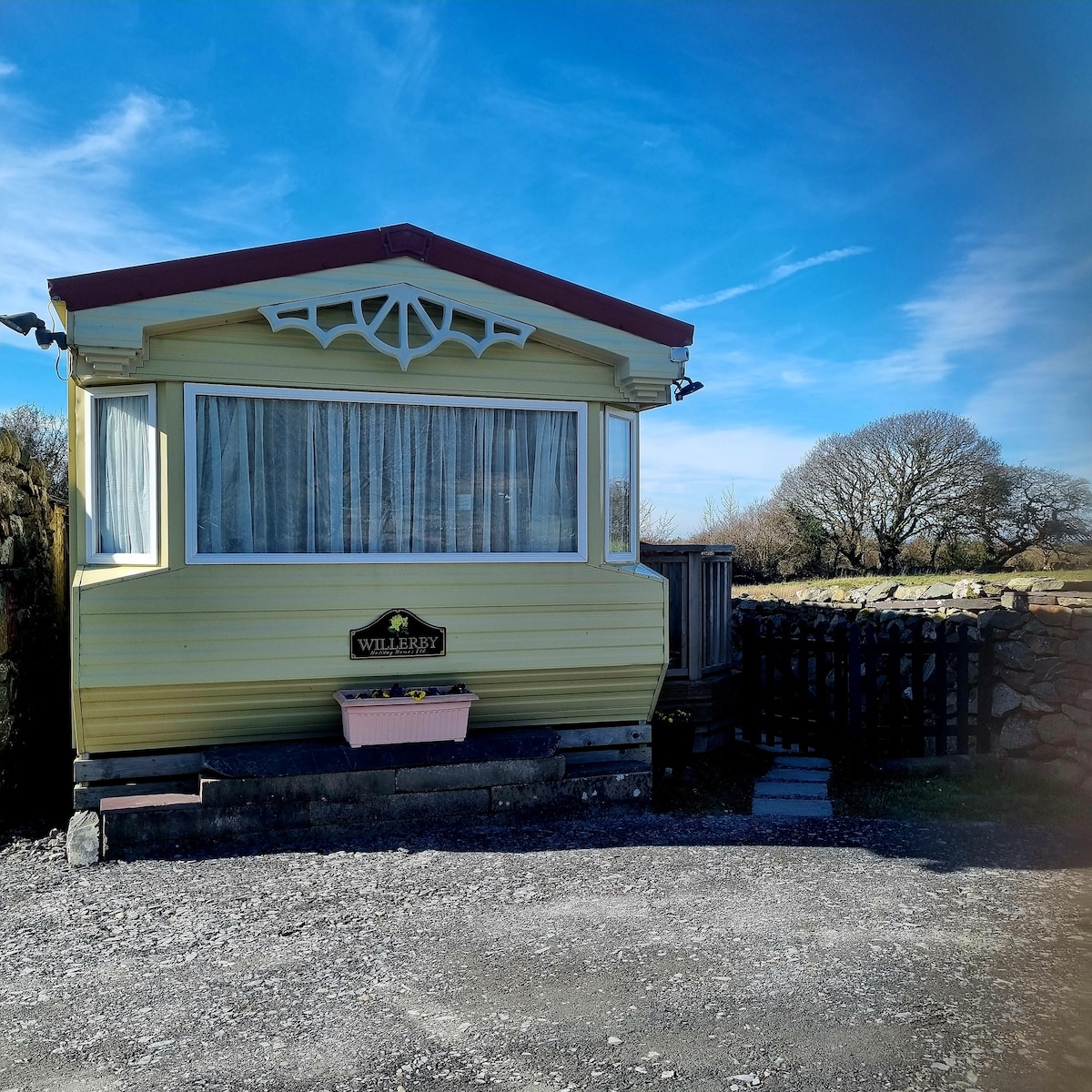 The exterior of a cozy yellow caravan is presented, featuring a large front window framed by lace curtains. The caravan is complemented by a decorative sign and a flower box underneath the window. The surrounding gravel area and a secure wooden fence enhance the property’s appeal.