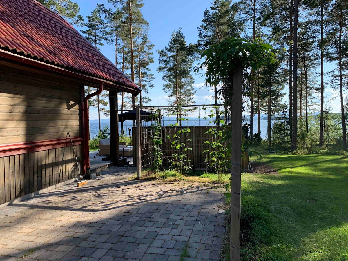 A wooden house with a red roof is viewed from a stone patio. Lush green surroundings and tall trees are visible, leading to a glimpse of a body of water in the distance. Outdoor seating and a pergola are suggested on the patio.