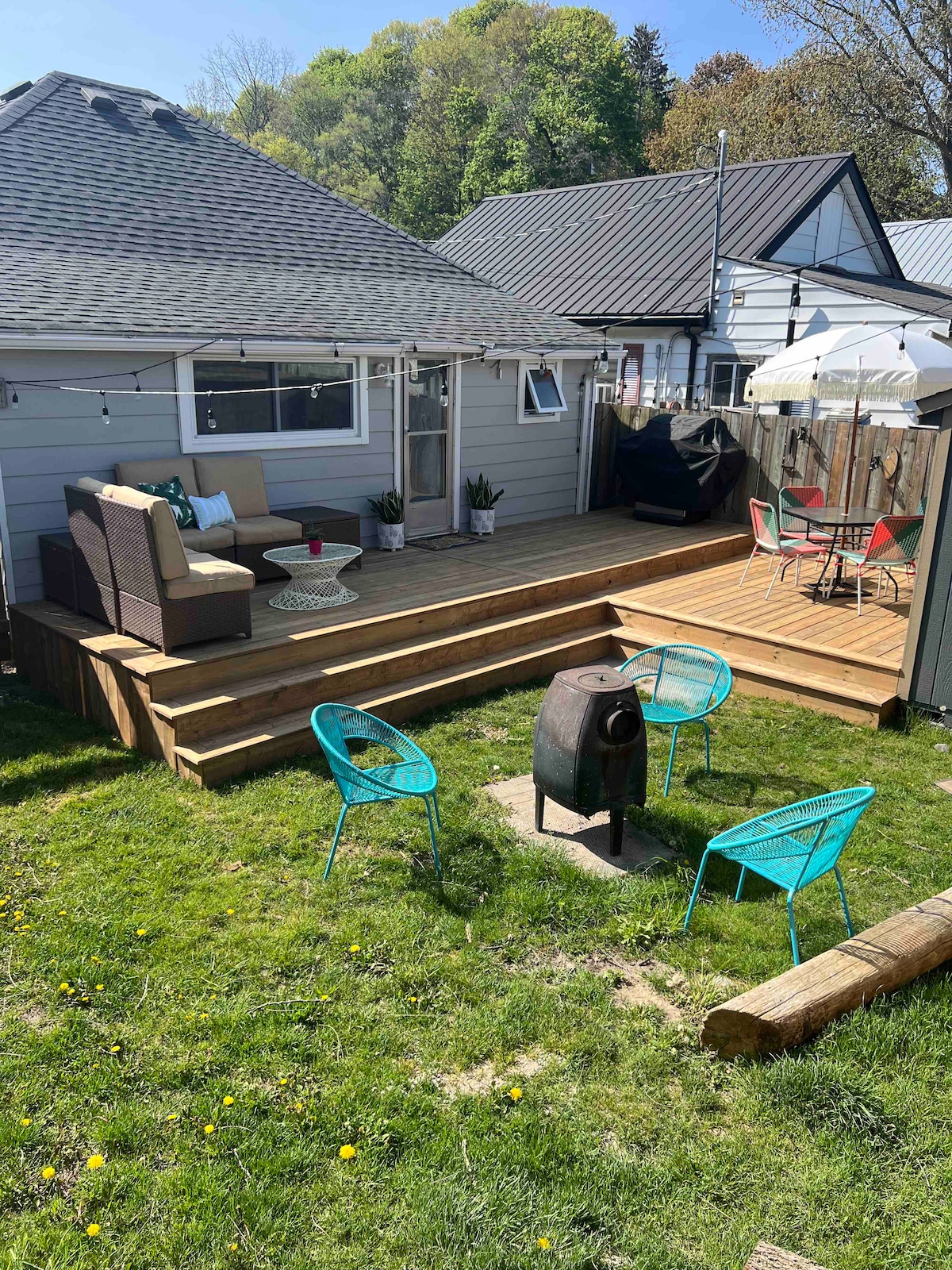 An outdoor deck area features a seating arrangement with a sectional sofa and a small table, surrounded by vibrant blue chairs. A circular fire pit sits on the grassy lawn, offering an inviting space for gatherings. A patio umbrella provides shade, enhancing the outdoor experience.